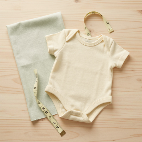 Petite barboteuse de naissance en coton biologique présentée avec un échantillon de tissu et un ruban de mesure sans marquage.
